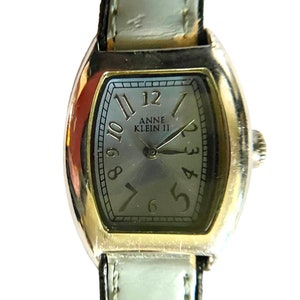 May include: A silver and gold toned watch with a white leather strap. The watch face is oval shaped and has black Roman numeral markings. The watch face also has the text "Anne Klein II" printed on it.