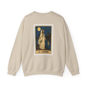 May include: A beige long-sleeve sweatshirt featuring a rectangular graphic print on the back. The print depicts a figure in a white dress standing by water under a moonlit sky, with the text LA LLORONA and the number 3 printed at the bottom.