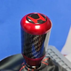 May include: A cylindrical gear shift knob featuring a carbon fibre centre section, a metallic red top cap with a red H logo on a black background, and a black leather shift boot with red stitching at the base of the gear shift assembly.