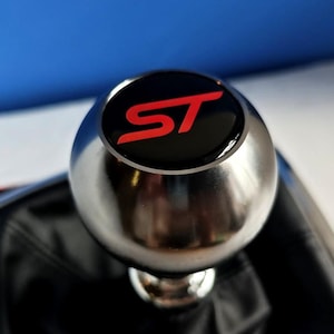 以下が含まれることがあります： 中央に赤い文字でSTとプリントされた黒い円形のトップインサートを備えた、球形のシルバーのシフトノブ。このノブは、目に見えるステッチが施された黒いレザー調のシフトブーツに取り付けられており、車両のインテリア用に設計されています。