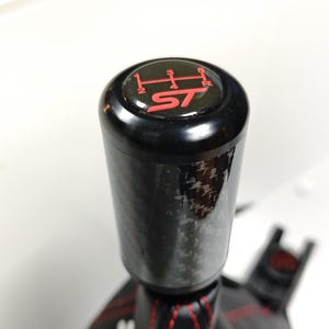 May include: A cylindrical gear shift knob featuring a black carbon fiber finish and a top cap displaying a red six-speed manual transmission pattern with the letters ST. The knob is attached to a black leather shift boot with red stitching.