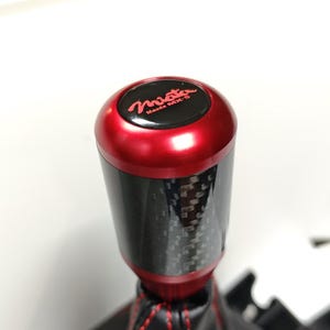 May include: A cylindrical gear shift knob featuring a red metallic top and a black carbon fibre body. The top cap displays the red text Miata Mazda MX-5. Attached to the base is a black leather shift boot with prominent red stitching.