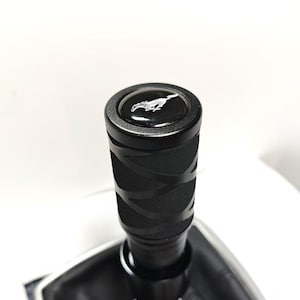 Pode incluir: Um manípulo de mudanças cilíndrico preto com um padrão de aderência texturizado e um inserto circular no topo exibindo um emblema de cavalo branco a galope. O manípulo está fixado a um fole de couro sintético preto com rebordo prateado.