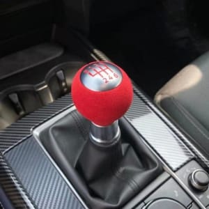 Op de afbeelding: Een bolvormige rode versnellingspookknop met een zilverkleurige bovenkant voorzien van een rood schakelpatroon voor een handgeschakelde zesversnellingsbak. De knop is gemonteerd op een zwarte lederen hoes in een middenconsole met carbonafwerking.