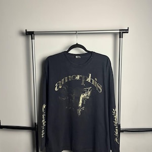 May include: A black long-sleeve t-shirt featuring the word "amorphis" in a light-colored, distressed font arched over a central graphic of a horned creature. The sleeves also display matching light-colored, distressed graphic patterns.