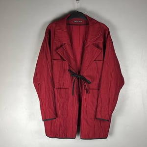 Pode incluir: Um casaco de manga comprida em vermelho escuro com textura enrugada, apresentando um colarinho largo, dois bolsos frontais grandes e um fecho de fita preta fina na cintura. A peça tem acabamentos pretos nos punhos e na bainha inferior.