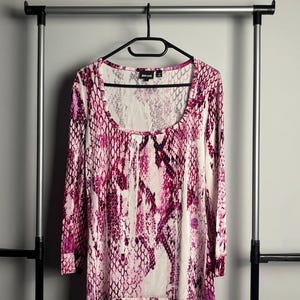 May include: A long-sleeve top featuring a vibrant pink and white snakeskin pattern throughout. The garment has a wide scoop neckline and a straight hemline. A black brand label is visible at the inner neck area of the shirt, which is displayed on a black hanger.