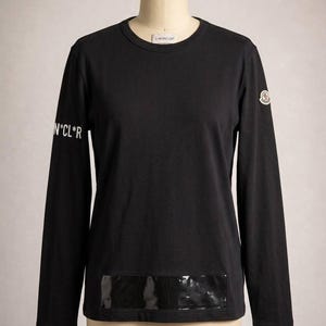 May include: A black long-sleeve crew neck shirt featuring the text N*CL*R printed in white on the right sleeve, a circular logo patch on the left sleeve, and a rectangular glossy black panel detail at the bottom hem on the front.