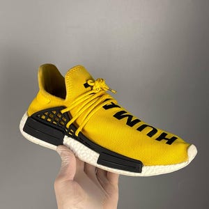 May include: A bright yellow sneaker with a knit upper featuring the black text HUMAN printed on the front. The shoe includes a white textured midsole, black plastic side cage overlays, matching yellow laces, and a black heel tab detail.