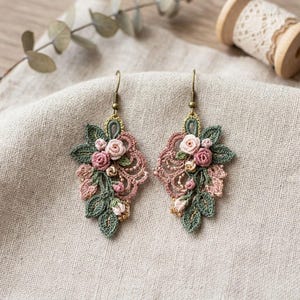 Könnte beinhalten: Ein Paar Ohrhänger mit aufwendigen Häkel-Blumenmotiven in Altrosa und Salbeigrün. Das Design umfasst kleine gestickte Rosen, zarte Blätter und goldfarbene Metallhaken zur Befestigung an den Ohrläppchen.
