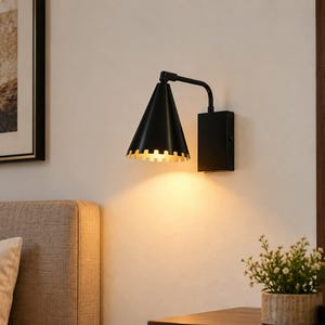 May include: A black wall-mounted lamp featuring a conical shade with a scalloped bottom edge. The interior of the shade is gold-toned, emitting a warm light downward. The fixture is attached to a rectangular black base mounted on the wall.