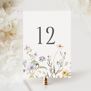 May include: A table number card with the number 12 in dark gray. The bottom of the card features a watercolor floral design with purple, yellow, and white flowers. The card is held by a copper-colored metal stand.