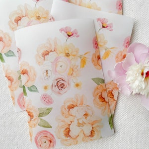 Op de afbeelding: Een stapel witte kaarten met een aquarel bloemenpatroon in tinten roze, perzik en geel. De kaarten liggen op een wit, getextureerd oppervlak.