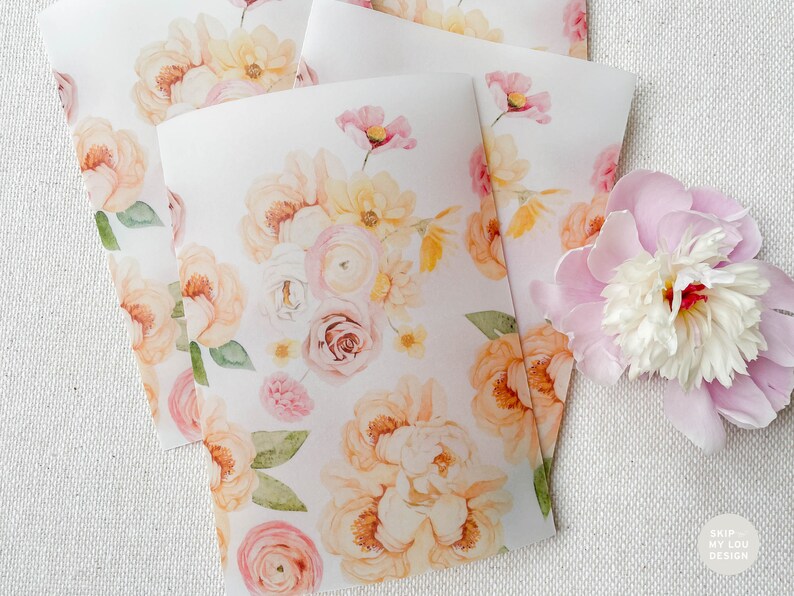 Op de afbeelding: Een stapel witte kaarten met een aquarel bloemenpatroon in tinten roze, perzik en geel. De kaarten liggen op een wit, getextureerd oppervlak.