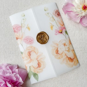 Op de afbeelding: Een witte envelop met een bloemenontwerp en een gouden waszegel. De bloemen zijn roze, perzik en geel. De envelop ligt op een beige stoffen achtergrond.
