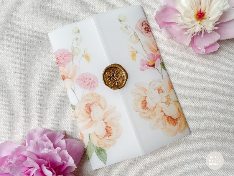 Op de afbeelding: Een witte envelop met een bloemenontwerp en een gouden waszegel. De bloemen zijn roze, perzik en geel. De envelop ligt op een beige stoffen achtergrond.