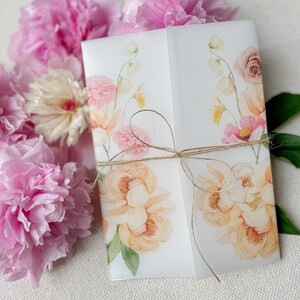 Op de afbeelding: Een witte uitnodigingskaart met een aquarel bloemenontwerp met roze, gele en groene bloemen. De kaart is vastgebonden met een bruin touwtje.