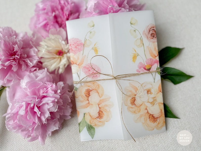 Op de afbeelding: Een witte uitnodigingskaart met een aquarel bloemenontwerp met roze, gele en groene bloemen. De kaart is vastgebonden met een bruin touwtje.