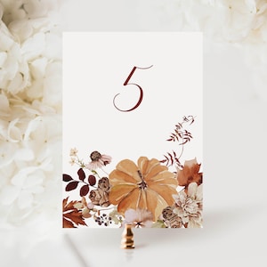 May include: A white card with a brown floral design and the number 5 printed in maroon. The card is on a gold stand.
