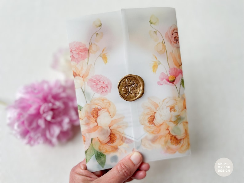 Op de afbeelding: Een witte envelop met een bloemenontwerp in tinten roze, perzik en groen. De envelop heeft een gouden waszegel met een bloemenontwerp.