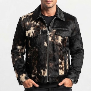 May include: A long-sleeve jacket featuring a textured cowhide pattern in black, brown, and white. The garment includes a pointed collar, a full-length silver-toned metal zip closure, and two chest pockets with flaps. The material has a visible fur texture.
