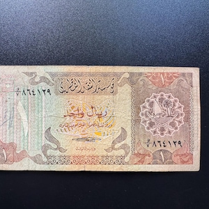 Puede incluir: Un billete rectangular de un riyal con intrincados patrones en tonos marrón, beige y verde. El billete muestra escritura árabe, el número uno en cifras árabes y occidentales, y un emblema circular que representa un barco dhow y una palmera.