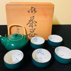 以下が含まれることがあります： 湾曲した竹製の持ち手が付いた深緑色の陶器製ティーポットと、お揃いの小さなティーボウル5客のセット。ボウルは内側が白く、繊細な青い花の模様が描かれています。セットは木製の収納箱に入っています。