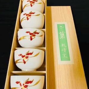 May include: A set of five white ceramic tea bowls arranged in a vertical wooden box. Each bowl features a hand-painted red orchid design with gold accents. The wooden box lid includes a green rectangular label with white Japanese calligraphy text.
