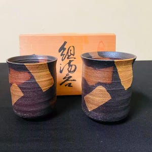 May include: Two cylindrical ceramic cups with a dark, textured finish featuring irregular patches of tan and brown. Behind the cups sits a light-colored wooden box with black Japanese calligraphy characters printed on the front surface.
