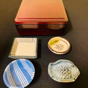 May include: A dark red rectangular wooden box positioned behind four small ceramic dishes. The collection includes a square dish with orange and blue stripes, a round dish with a green accent and black symbol, a blue striped bowl, and a fish-shaped dish.