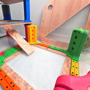 Peut inclure: Un ensemble de construction modulaire composé de connecteurs en plastique vert vif, orange, jaune et rouge fixés sur des panneaux de particules. La structure comprend une rampe en bois, une feuille acrylique transparente et un panneau en bois avec une fenêtre grillagée.