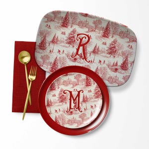 May include: A red and white serving tray and plate set with a winter scene toile pattern. The tray and plate feature the letters "R" and "M" respectively. Gold-colored fork and spoon are on a red napkin.