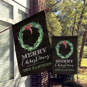 May include: A dark wooden Christmas flag with a watercolor wreath and a red bow. The words "MERRY CHRISTMAS" are written in white script, with "THE CARTERS" below in green.