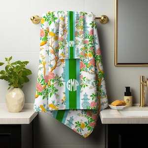 May include: White bath towels with a floral and pagoda design, accented with pink and yellow flowers, green leaves, and a green stripe with the initials "GMP". The towels are hanging on a gold towel rack in a bathroom setting.
