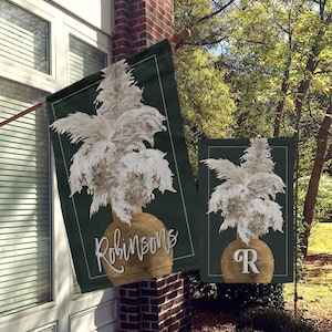 May include: A pair of green garden flags with a white pampas grass design. The larger flag has the name "Robinson's" written in gold script on the vase. The smaller flag has the letter "R" in gold on the vase.