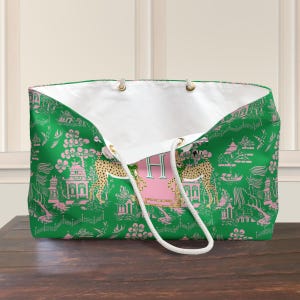 May include: A large tote bag with a green and pink Asian-inspired pattern. The bag features a white interior and rope handles. A pink rectangle with the letter "H" is centered on the front. The bag's design includes pagodas, flowers, and cheetahs.