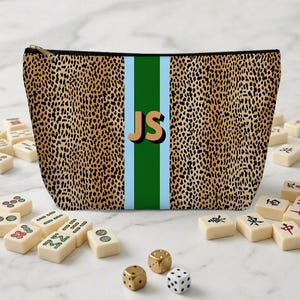 May include: A leopard print cosmetic bag with a vertical green stripe and light blue borders. The initials "JS" are in the center. Mahjong tiles and dice are scattered around the bag.