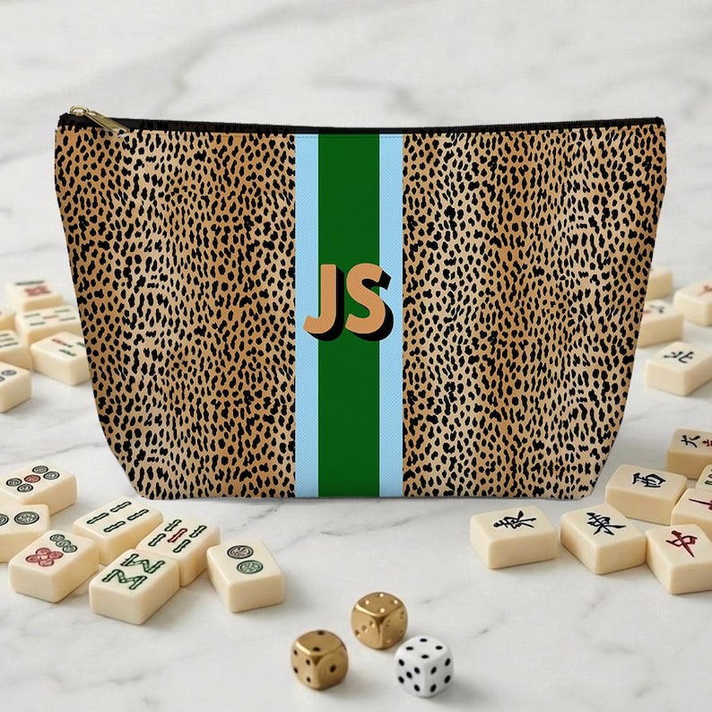 May include: A leopard print cosmetic bag with a vertical green stripe and light blue borders. The initials "JS" are in the center. Mahjong tiles and dice are scattered around the bag.