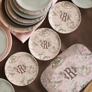 May include: A collection of pink and brown decorative plates and a serving tray. The plates and tray feature a landscape design with trees, buildings, and the letter "R". The plates are stacked and arranged on a brown surface.
