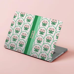 Puede incluir: Ordenador portátil con un estampado floral. El diseño presenta flores rosas y hojas verdes sobre un fondo blanco. Una franja verde con la letra "H" está en el lado izquierdo del portátil.
