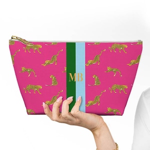 May include: A pink cosmetic bag with a gold zipper and a repeating pattern of yellow and black leopards. The bag has a green and light blue stripe with the gold letters "MB".