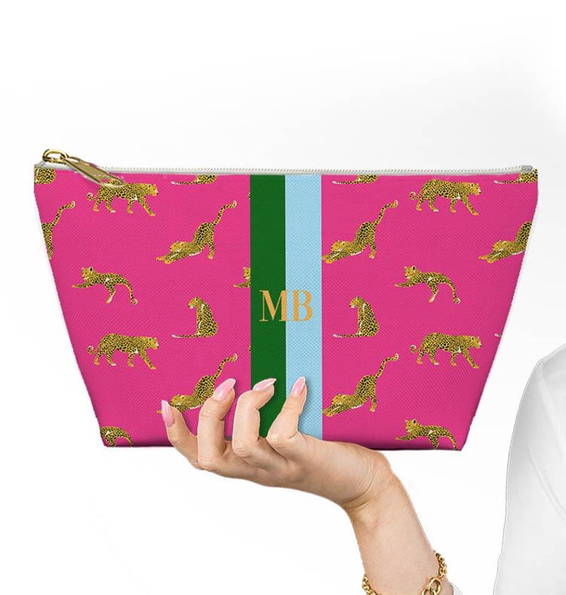 May include: A pink cosmetic bag with a gold zipper and a repeating pattern of yellow and black leopards. The bag has a green and light blue stripe with the gold letters "MB".