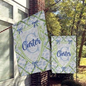 May include: Two garden flags with a blue and green design. The flags feature a white shield with a blue letter "C" and the name "Carter" in blue script. The flags are hanging on a brick wall.