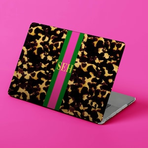 May include: Laptop skin with a tortoiseshell pattern, a pink and green stripe, and the initials "SEH" in gold. The laptop is on a bright pink background.