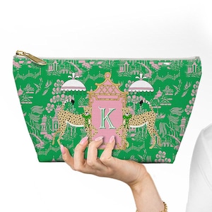 May include: A green cosmetic bag with a pink and white design featuring cheetahs, pagodas, and the letter "K". The bag has a gold zipper and is held by a hand. The bag's pattern is a toile design.