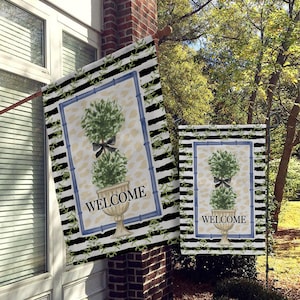 May include: Two welcome flags with a black and white striped background, green topiary plants in white pots, and the word "Welcome" in blue lettering.
