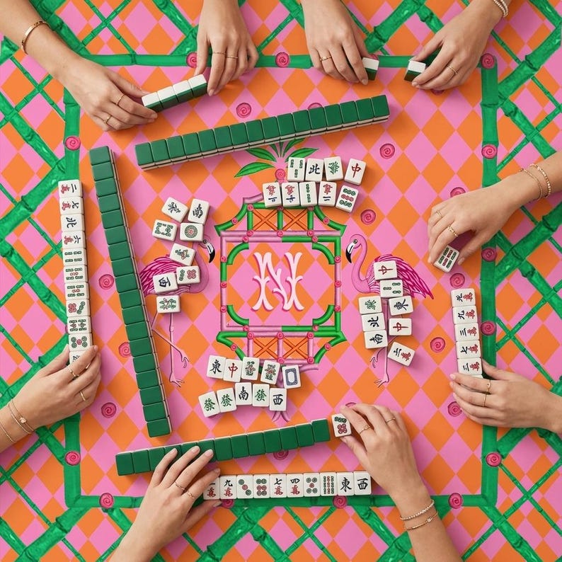 May include: A mahjong set featuring white tiles with black, red, and green characters and symbols, arranged on a pink and orange diamond-patterned mat with green bamboo borders and pink flamingo illustrations surrounding a central letter N.