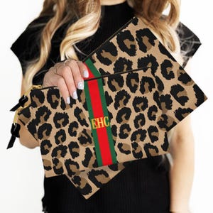 May include: Two leopard print clutches with a red and green stripe down the center. The clutches have gold zippers and the initials "EHC" in gold. The background is a light color.