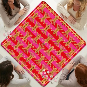 K&ouml;nnte beinhalten: Eine quadratische Spielmatte mit einem sich wiederholenden Muster aus orangefarbenen und schwarzen Tigern auf einem Hintergrund aus abwechselnden diagonalen Streifen in Pink und Rot. Die Matte hat einen d&uuml;nnen goldenen Rand und die Buchstaben SHN.