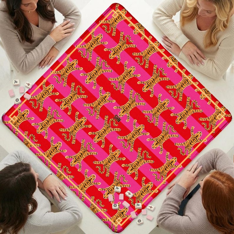 K&ouml;nnte beinhalten: Eine quadratische Spielmatte mit einem sich wiederholenden Muster aus orangefarbenen und schwarzen Tigern auf einem Hintergrund aus abwechselnden diagonalen Streifen in Pink und Rot. Die Matte hat einen d&uuml;nnen goldenen Rand und die Buchstaben SHN.